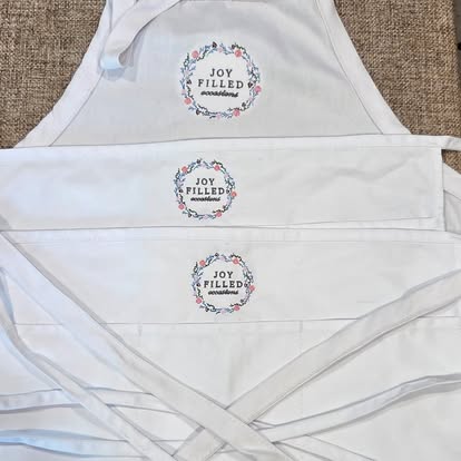 Three white aprons embroidered with the Joy Filled Occasions logo