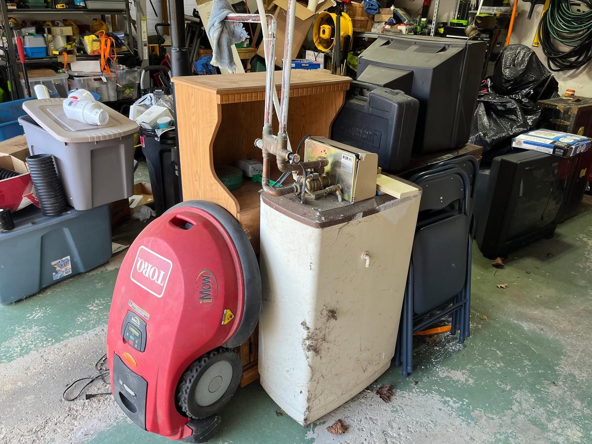 A packed garage mid-cleanout: mower, stained water heater, cabinet, tube televisions and folding chairs