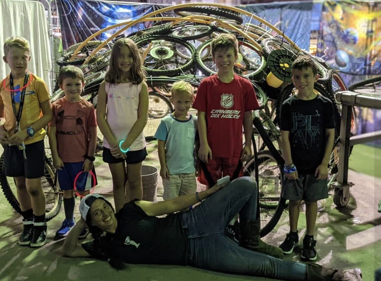 Mary lying on the ground grinning in front of six kids, in front of a large dome sculpture built from salvaged bicycle wheels and frames