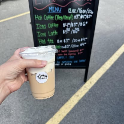 An iced latte in hand in front of the Little A's chalkboard menu